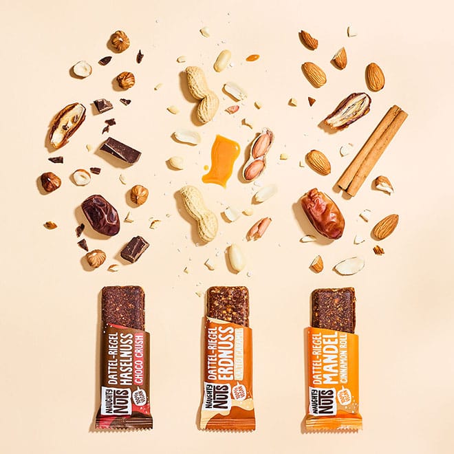 Drei Naughty Nois Dattel-Riegel: Haselnuss, Erdnuss, Mandel, umgeben von Nüssen, Datteln, Schokolade und Zimt